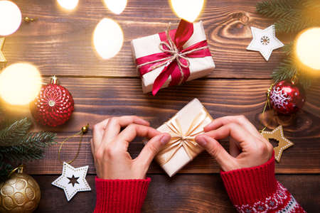 Women's hands in knitted sweater with ornament hold a box with Christmas gift in their palms on the table among the decor. New year, packaging and preparation of gifts, branches of fir, decorationの写真素材