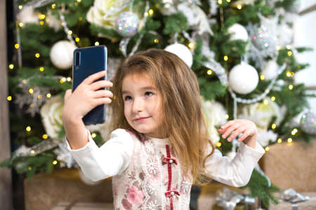 Little Caucasian girl in beautiful dress on background of Christmas tree takes photos of herself with smartphone, looks at screen, smiles, poses and makes faces. Video communication, new year greetingsの写真素材