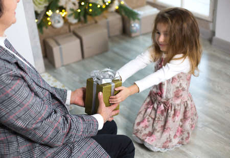 Grandma gives her granddaughter a gift for Christmas. The girl stretches out her hands to the box with the bow. New year, family holiday, communication of generationsの写真素材