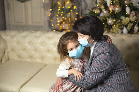 Grandmother and granddaughter on the sofa in the living room with Christmas decor hugging in medical masks on their faces. A family holiday during the outbreak of coronavirus and disease. New Yearの写真素材