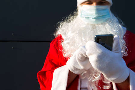 Santa Claus in a medical mask with a smartphone. Online greetings, ordering services for Christmas and new year. Social distance in the coronavirus epidemic, a new reality. Copy spaceの写真素材