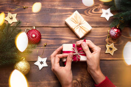Women's hands in knitted sweater with ornament hold a box with Christmas gift in their palms on the table among the decor. New year, packaging and preparation of gifts, branches of fir, decorationの写真素材