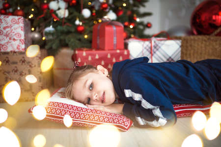 A little girl in home clothes is lying under the Christmas tree near the boxes with gifts on the pillow and dreams. Lights of garlands, the night before Christmas, new year.の写真素材
