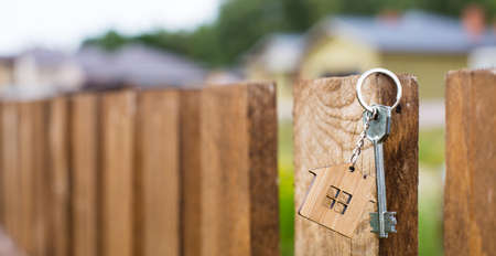 Wooden pendant of a house and key. Background of fence and cottage. Dream of home, building, design, delivery of the project, moving to a new house, mortgage, rent and purchase real estate. Copy spaceの写真素材
