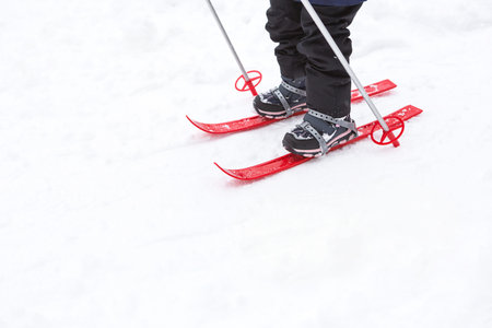 Children's feet in red plastic skis with sticks go through the snow from a slide-a winter sport, family entertainment in the open air. A little girl glides down the slope from an early age. Copy spaceの写真素材