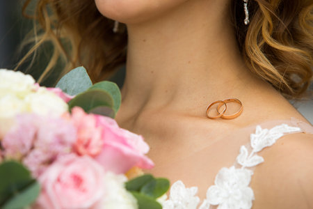 A pair of wedding rings of the bride and groom on the shoulder of the future wife in a white dress. Wedding accessories, ceremony, jewelry, Bridal bouquet of pink roses. Space for textの写真素材