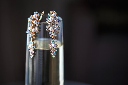 Large gold earrings with pearls and crystals on a clear glass of champagne. Morning and gathering of the bride, costume jewelry for the wedding celebration. Jewelry for the holiday. Black background, space for textの写真素材