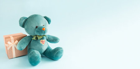 Turquoise soft teddy bear with an embroidered heart holds a gift box and a bow on a blue background. Children's toy. Love, a gift to holiday, a declaration of love, Valentine's day. Copy spaceの写真素材