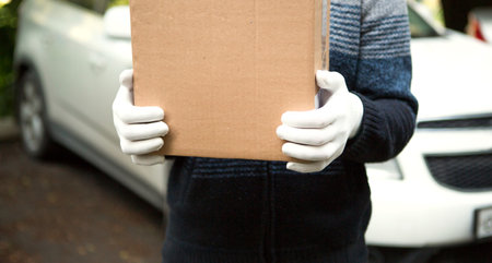 A cardboard box with space for text in the hands of a male courier in white gloves. Courier on the background of a white car. Contactless home delivery service for online orders. Mockupの写真素材