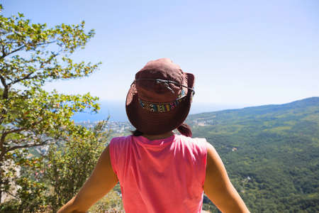 Woman in a hat looks at the panoramic view from the mountain to the sea and the forest. Tourist, trekking, travel. Active ecotourism, healthy lifestyle, adventureの写真素材