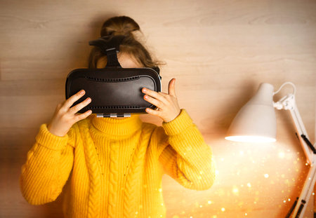A girl in virtual reality glasses sits near a lamp with a magic light. Yellow sweater, technology,...