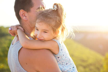 The father and daughter hug tightly, the girl holds the father by the neck, the father - in his arms. Father's Day, strong feelings, family. Summer, sunshine, greenery, mountains, travelの写真素材