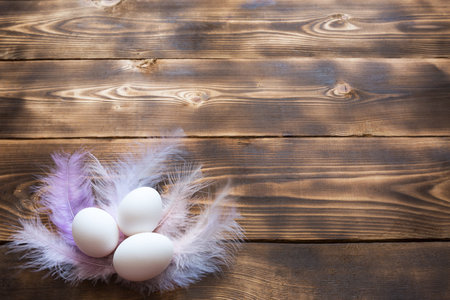 White chicken eggs on a wooden table in a nest of delicate pastel-colored feathers. Easter village...