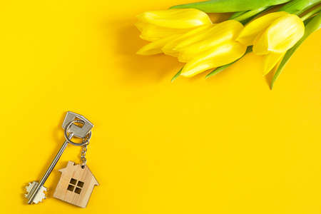 Key ring in the shape of wooden house with key on yellow background and spring tulips. Building, design, project, moving to new home, mortgage, rent and purchase real estate, summer offer. Copy spaceの写真素材
