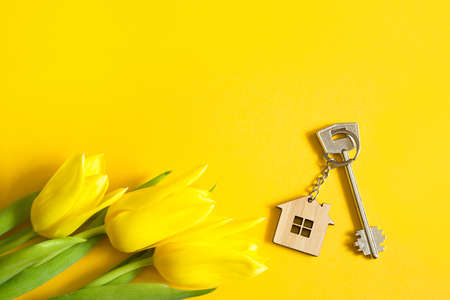 Key ring in the shape of wooden house with key on yellow background and spring tulips. Building, design, project, moving to new home, mortgage, rent and purchase real estate, summer offer. Copy spaceの写真素材