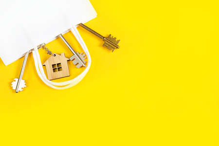 Key ring in the shape of wooden house with key on a yellow background in a white paper gift wrapping bag. Mortgage, rent and purchase real estate, beneficial offer for construction. Copy spaceの写真素材