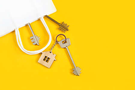 Key ring in the shape of wooden house with key on a yellow background in a white paper gift wrapping bag. Mortgage, rent and purchase real estate, beneficial offer for construction. Copy spaceの写真素材