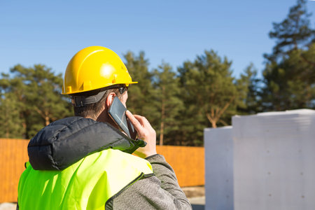 The builder talking on the phone. Architect in a protective yellow helmet and a signal vest stands...