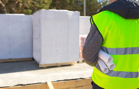 The project architect's back is on the construction site of a house with the foundation laid and the...