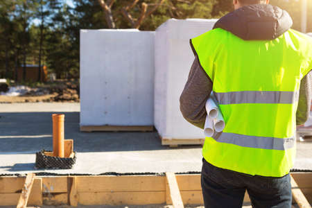 The project architect's back is on the construction site of a house with the foundation laid and the blocks delivered. Construction worker in a protective yellow helmet and a signal vest. Mock upの写真素材