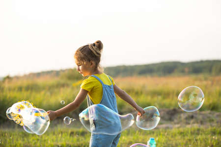 A girl in a denim jumpsuit blows soap bubbles in the summer in a field at sunset. International Children's Day, happy child, outdoor activities. Summer background. Healthy and eco-friendly lifestyleの写真素材