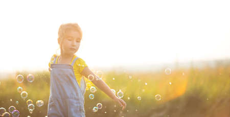 A girl in a denim jumpsuit blows soap bubbles in the summer in a field at sunset. International Children's Day, happy child, outdoor activities. Summer background. Healthy and eco-friendly lifestyleの写真素材