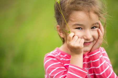 Little girl in a coral striped T-shirt on a green background in a field holds her face in her hands and smiles slyly. Children's Day, happy child, environmental and nature protection, insect repellentの写真素材