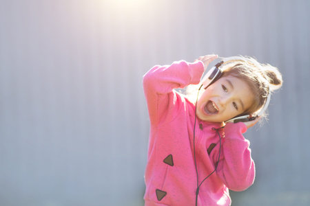 A cheerful girl in headphones and a pink hoodie listens to music and laughs. Outdoor, gray backgroundの写真素材