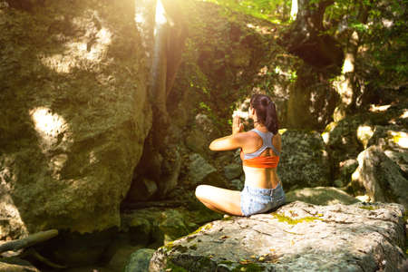 A woman sits in a lotus field on a large rock among the rocks in the open air and meditates, enjoys the unity with nature, listens to the silence and sounds of the forest. ecologyの写真素材