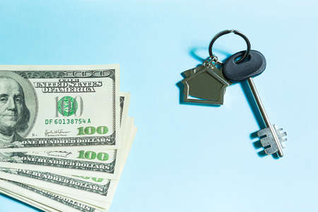 Home key with a keychain on a stack of $ 100 bills on a blue background. Purchase of an apartment, house, real estate, business, mortgage and housing loan from bank, savings, cash, moving. Copy spaceの写真素材