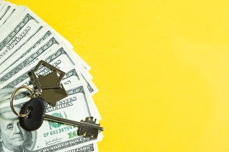 Home key with a keychain on a stack of $ 100 bills on yellow background. Purchase of an apartment, house, real estate, business, mortgage and housing loan from bank, savings, cash, moving. Copy spaceの写真素材