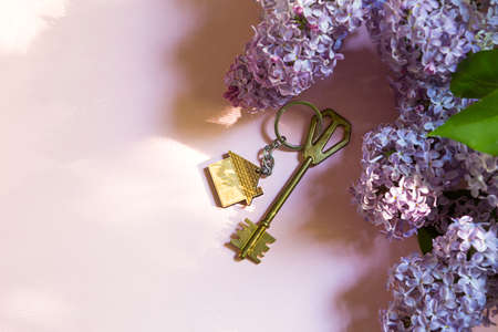 House key with keychain on pink spring background and lilac branches. Summer holiday home, booking cottage in the countryside, moving to new home, mortgage, rent and purchase real estate. Copy spaceの写真素材