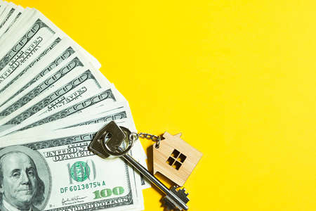 Home key with a keychain on a stack of $ 100 bills on yellow background. Purchase of an apartment, house, real estate, business, mortgage and housing loan from bank, savings, cash, moving. Copy spaceの写真素材