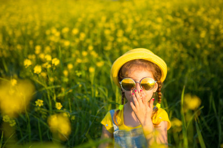 A girl in a yellow hat and round glasses looks at the sun in a summer blooming field. Summer time,...