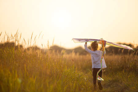A girl runs into a field with a kite, learns to launch it. Outdoor entertainment in summer, nature and fresh air. Childhood, freedom and carelessness. A child with wings is a dream and a hope.の写真素材