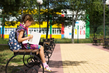 Girl with a backpack sitting on a bench and reading a book near the school. Back to school, lesson schedule, a diary with grades. Education, primary school classes, September 1の写真素材