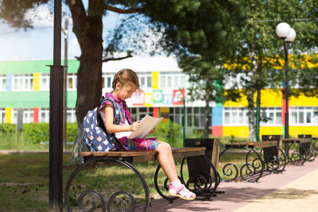 Girl with a backpack sitting on a bench and reading a book near the school. Back to school, lesson schedule, a diary with grades. September 1. The inscription on the building in Russian "School"の写真素材