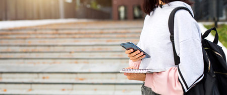 A female student with a black backpack holds a smartphone and Notepad on the threshold of the University. Higher education, the beginning of the school year, return to school, gadgets. Copyspaceの写真素材
