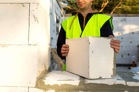 The builder holds a block of cellular concrete in his hands - the masonry of the walls of the house. Construction workers in protective clothing-a hardhat and a reflective vest.の写真素材