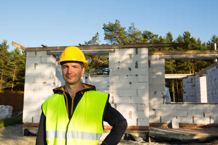 Project architect stands near construction site of a house with the walls are made of a porous concrete block and a brick. Construction worker in a protective yellow hardhat and a reflective vest.の写真素材