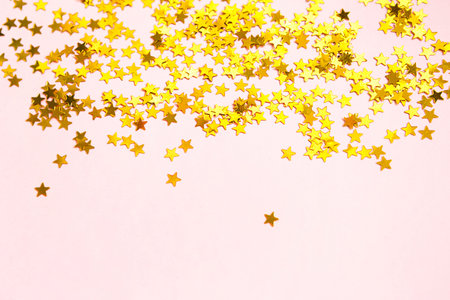 Gold sequins in the shape of stars shimmer on a pink background. Backgrounds with copyspace for a...