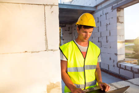 Construction engineer on the construction site of a house made of porous concrete blocks works at a computer in a reflective safety vest and hardhat. Design, construction, drawings project, checkの写真素材