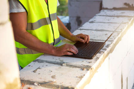 Construction engineer on the construction site of a house made of porous concrete blocks works at a computer in a reflective safety vest and hardhat. Design, construction, drawings project, checkの写真素材
