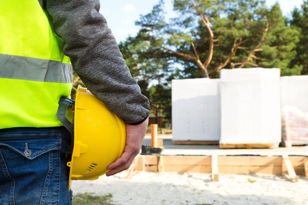 Builder holds in hand a yellow protective hardhat on the construction site with the foundation and walls of the house made of cellular concrete blocks and construction materials. Work safety, mockupの写真素材
