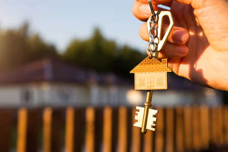 Key ring with keychain house in hand. Background of fence and cottage. Moving to a new home, mortgage, buying real estate, renting and booking housing, dream of living in the village. Copy spaceの写真素材