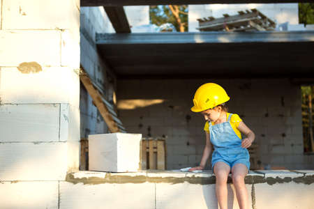 Little girl in a yellow hardhat is playing builder on the construction site of her future home. Expectation of moving, choosing a profession, children's education. Future profession, family business.の写真素材