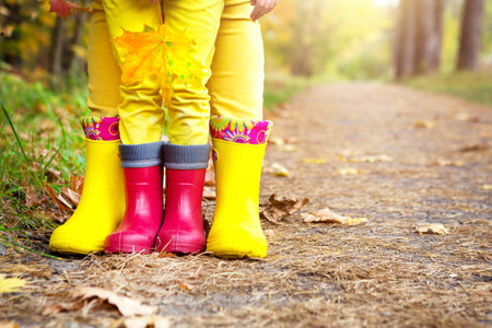 Feet in red and yellow rubber boots of a mother and daughter in the autumn forest. Seasonality, seasons, fallen dry maple leaves, family walk, feeling of love and care, parenthoodの写真素材