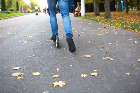 The legs of a man in jeans and sneakers on a scooter in the park in autumn with fallen dry yellow...