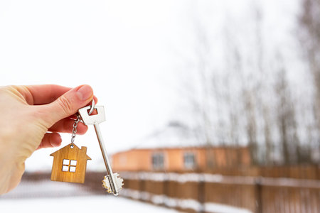 Hand with a Key of the house in winter in the snow. Background of fence and cottage. Give a gift for New Year, Christmas. Moving to new house, mortgage, rent and purchase real estate. Open the door.の写真素材