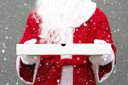 White pizza box in the hands of Santa Claus in white mittens, with a beard, in a red coat. Christmas fast food delivery. New year's eve promotion. Work on public holidays catering. Copy space, mock upの写真素材